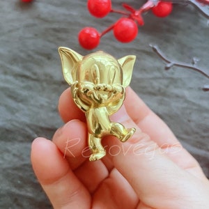 May include: A gold-coloured figurine of a cartoon cat with its arms raised. The cat is standing on its hind legs and has a smiling expression. The figurine is held in a hand, with red berries in the background.