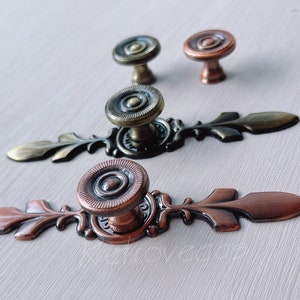 May include: Three antique-style cabinet knobs with ornate metalwork. Two of the knobs are copper-toned, and one is a darker bronze colour. Each knob has a decorative, circular design with a raised centre.