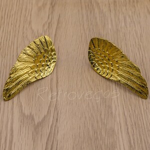 Peut inclure: Une paire de boucles d'oreilles dorées en forme d'ailes d'ange. Les ailes ont un design texturé et plumeux.