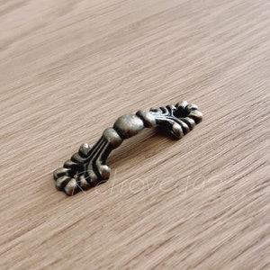 May include: A small, ornate, antique-style drawer pull with a decorative, scrolled design. The pull is made of metal and has a bronze finish.