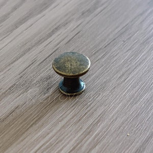 Könnte beinhalten: Ein kleiner Schrankknauf im antiken Stil mit einer runden, bronzefarbenen Oberseite und einem dunklen Sockel. Der Knauf ist auf einer hellbraunen Holzoberfläche platziert und bietet eine Vintage-Ästhetik.