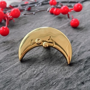 May include: A gold crescent moon shaped pin with two small circles on the top. The pin is on a grey textured surface.