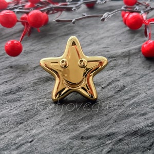 May include: A gold-colored star-shaped pin with a smiling face. The pin is on a gray textured surface.