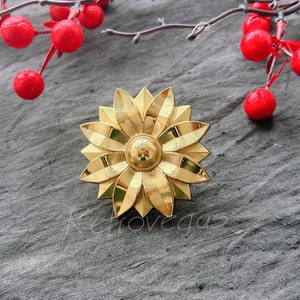 May include: A gold-coloured metal flower-shaped decorative piece with a raised centre. The piece has a detailed design and is reminiscent of a stylised flower.