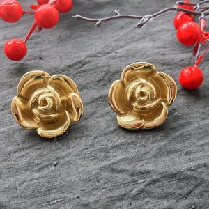 May include: A pair of gold-coloured rose-shaped stud earrings. The earrings are highly polished and reflect light. They are set against a dark grey textured surface, with red berries and branches in the background. Jewellery.