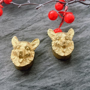 May include: Two gold-colored, detailed, three-dimensional dog head figurines. The figurines are on a gray textured surface.
