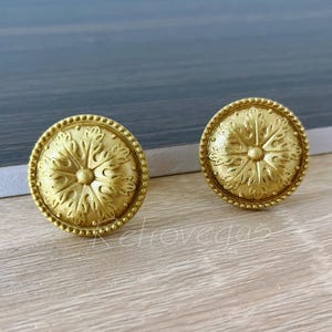 Puede incluir: Dos pomos de armario redondos dorados con un diseño floral. Cada pomo tiene un patrón detallado en relieve con un borde de cuentas. Los pomos se muestran sobre una superficie de madera clara.