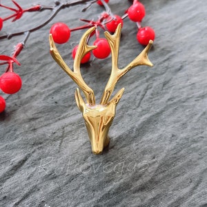 May include: A gold-coloured deer head ornament with a simple design. The ornament is on a grey background with red berries.
