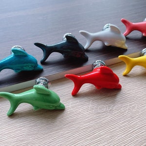 Peut inclure: Ensemble de sept poignées de tiroir en céramique en forme de dauphin dans différentes couleurs, notamment turquoise, vert, noir, blanc, rouge, jaune et rose. Chaque poignée présente un motif de dauphin avec un bouton en métal argenté.