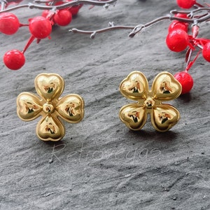 May include: A pair of gold-tone earrings shaped like four-leaf clovers. The earrings are on a grey textured surface.