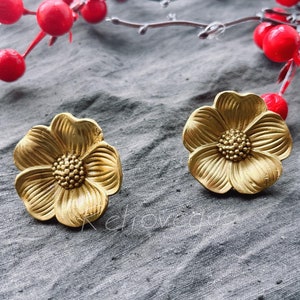 Puede incluir: Un par de pendientes de color dorado en forma de flor. Los pendientes están hechos de metal y tienen una superficie texturizada.