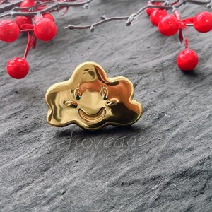 Op de afbeelding: Een gouden metalen pin in de vorm van een wolk met een glimlach.