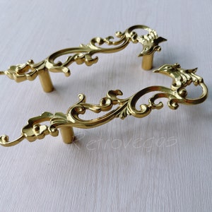 May include: Two ornate gold-toned cabinet pulls with a decorative scroll design. The pulls have a classic, vintage style.