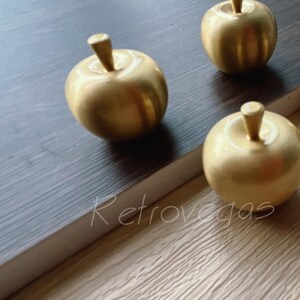 Op de afbeelding: Vier goudkleurige metalen decoratieve voorwerpen in de vorm van een appel op een donker houten oppervlak.