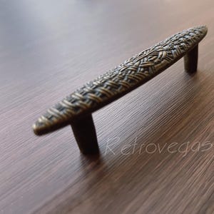 May include: A vintage-style,  brass cabinet pull with a woven pattern. The pull is shaped like a slightly curved, elongated oval.