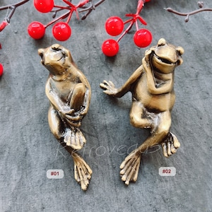 May include: Two gold-coloured frog figurines with smiling faces. The frog on the left is holding its foot, while the frog on the right is resting its chin on its hand. Both frogs have detailed features and are sitting on a grey textured surface.