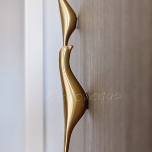 Puede incluir: Dos tiradores de puerta de armario en forma de pájaro dorados montados en una superficie de madera.