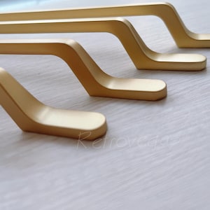 May include: Four gold-colored cabinet pulls with a curved, modern design. The pulls are arranged in a row, with the top pull slightly offset from the others.