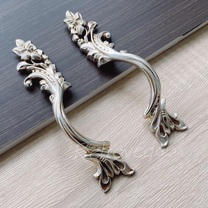 Può includere: Due tiretti per cassetti in argento decorati con un motivo floreale. I tiretti hanno una forma curva e sono progettati per essere utilizzati su armadi o mobili.