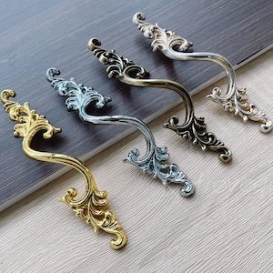 Op de afbeelding: Vijf sierlijke gouden en zilveren ladegrepen met ingewikkelde bloemmotieven. De grepen zijn in een rij op een lichtbruin houten oppervlak gerangschikt.