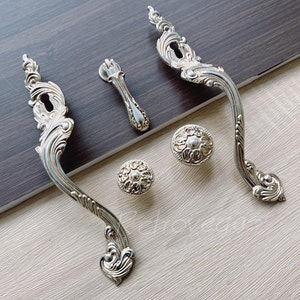 May include: A collection of silver-coloured cabinet hardware with an ornate design. The set includes two long handles, a small pull, and two round knobs. The hardware features detailed scrollwork and keyhole designs, suitable for vintage furniture.