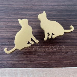 May include: Two gold-coloured cat-shaped decorative objects. The cats are in a sitting position with their tails curled. The objects are on a dark brown wooden surface. The word "retrovegas" is printed in white.