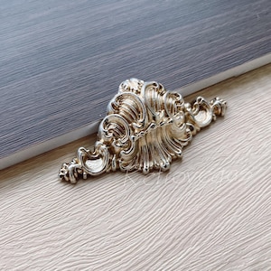 Op de afbeelding: Een gouden decoratieve kastbeslag met een gedetailleerd, sierlijk ontwerp. De greep heeft een schelpvorm met ingewikkelde krulwerk en een verhoogd midden.