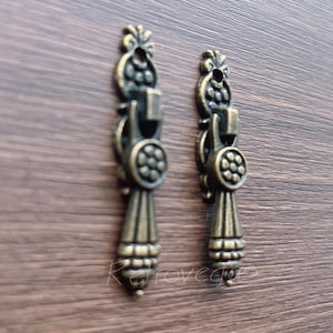 Puede incluir: Dos tiradores de cajón antiguos de color bronce con diseños florales y geométricos ornamentados. Cada tirador presenta una parte superior decorativa, un centro circular con un patrón de flores y una forma de lágrima estilizada en la parte inferior. Los tiradores están sobre un fondo de vetas de madera oscura.