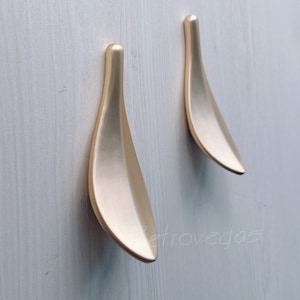 May include: Two gold leaf-shaped cabinet pulls, mounted on a white wood surface.