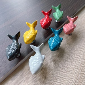 Könnte beinhalten: Set mit 7 Keramik-Fisch-Türknöpfen in verschiedenen Farben, darunter Blau, Grün, Rot, Gelb, Rosa, Grau und Weiß. Die Fische sind alle nach rechts gerichtet und haben ein 3D-Design.