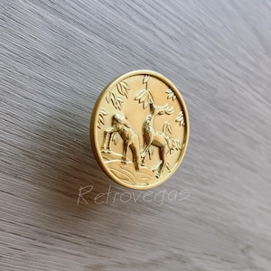May include: Gold-toned metal cabinet knob featuring a raised design of two deer standing in a woodland scene. The knob is round and has a smooth finish.