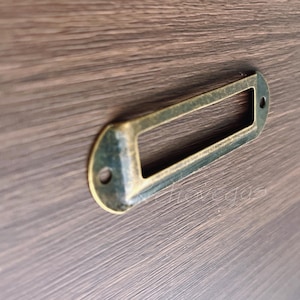May include: A brass drawer pull with a rectangular label holder. The pull has a worn, antique finish.