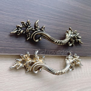 Peut inclure: Deux poignées de tiroir ornées de couleur or avec un motif floral décoratif. Les poignées sont en métal et ont une forme incurvée.