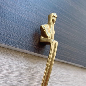May include: Gold-toned cabinet hardware in the shape of a seated figure with its chin resting on its hand. The figure is stylized and abstract.