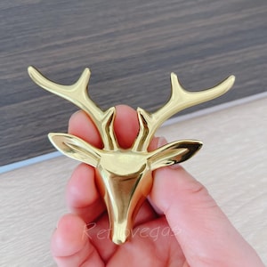 May include: A gold-colored deer head sculpture with detailed antlers and a polished finish. The sculpture is held in a hand, showcasing its size and design. The word "Retrovegas" is visible in the bottom left corner.