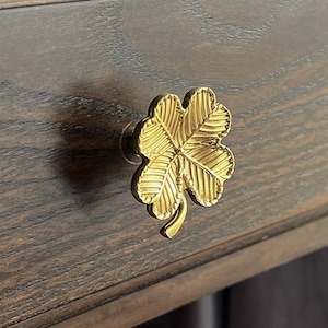 Puede incluir: Un tirador de cajón de trébol de cuatro hojas de color dorado con una superficie texturizada.