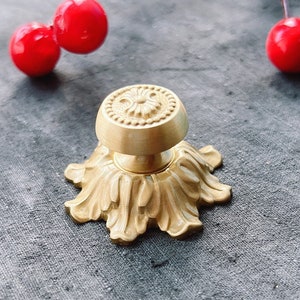 May include: A gold-toned metal door knob with a decorative floral design. The knob has a round top and a base with a scalloped edge.