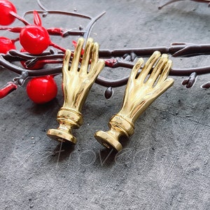 Puede incluir: Dos piezas decorativas doradas en forma de mano con una superficie texturizada. Las piezas están sobre un fondo gris con bayas rojas.