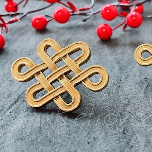 Può includere: Due nodi decorativi in metallo dorato, noti anche come nodo cinese o nodo portafortuna, sono mostrati su una superficie grigia testurizzata. I nodi sono intrecciati in modo intricato e hanno un design tradizionale.