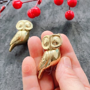 May include: Two gold owl figurines. One owl is sitting on a hand, the other is on a table. The owls have large eyes and detailed wings.