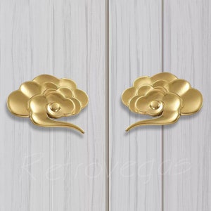 Puede incluir: Dos tiradores de puerta en forma de nube de color dorado con un diseño de espiral. Los tiradores están montados en una superficie de madera blanca.