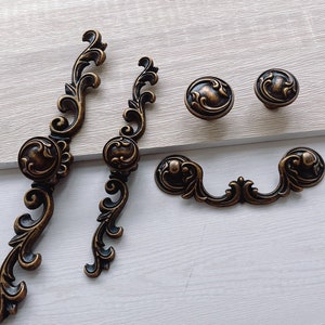 以下が含まれることがあります： 真鍮仕上げのアンティーク調のキャビネット金具プルが5つ。2つは、花柄の装飾が施された長いプル。3つは、花柄の丸いノブです。