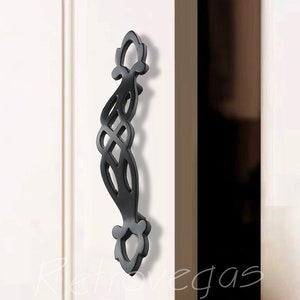 May include: Black metal cabinet door pull with a decorative, intricate design. The pull has a long, narrow shape with a curved top and bottom.