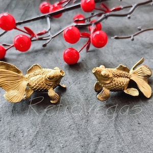 May include: Two gold-colored metal goldfish figurines. The figurines are detailed with scales and fins. They are sitting on a gray fabric surface.