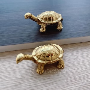 Könnte beinhalten: Zwei goldfarbene Schildkröten-Figuren. Die Schildkröten sind mit strukturierten Schalen detailliert und haben ein realistisches Aussehen.