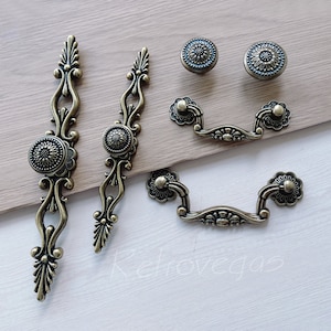 Könnte beinhalten: Satz von sechs antiken goldfarbenen Schrankbeschlägen. Das Set enthält zwei lange verzierte Griffe, zwei kleinere verzierte Griffe und zwei verzierte Schubladengriffe.