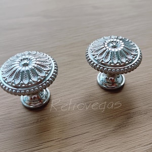 May include: Two silver cabinet knobs with a decorative floral design. The knobs have a round base and a raised, ornate top.