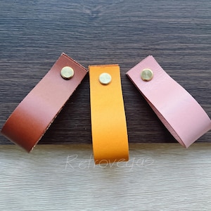 Könnte beinhalten: Drei Leder-Schubladengriffe in Braun, Orange und Rosa, jeweils mit einer Messingniete. Die Griffe sind auf einer dunklen Holzoberfläche angeordnet. Geeignet für Schrank- oder Schubladenbeschläge. Ca. 10 cm lang.