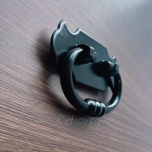 May include: Black metal drawer pull with a decorative, ornate design. The pull has a ring-shaped handle and a rectangular plate.