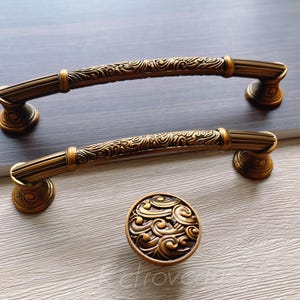 Op de afbeelding: Twee sierlijke, goudkleurige kastdeurgrepen met een decoratief golfpatroon. De grepen worden getoond op een lichtbruin oppervlak. Een bijpassende decoratieve knop wordt ook getoond.